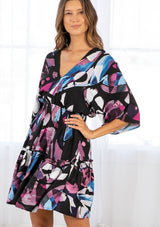 [Color: Black/Plum] A model wearing a black and purple bohemian watercolor print mini dress. With half length kimono sleeves, a smocked elastic waist, tiered skirt, and open tie back detail. 