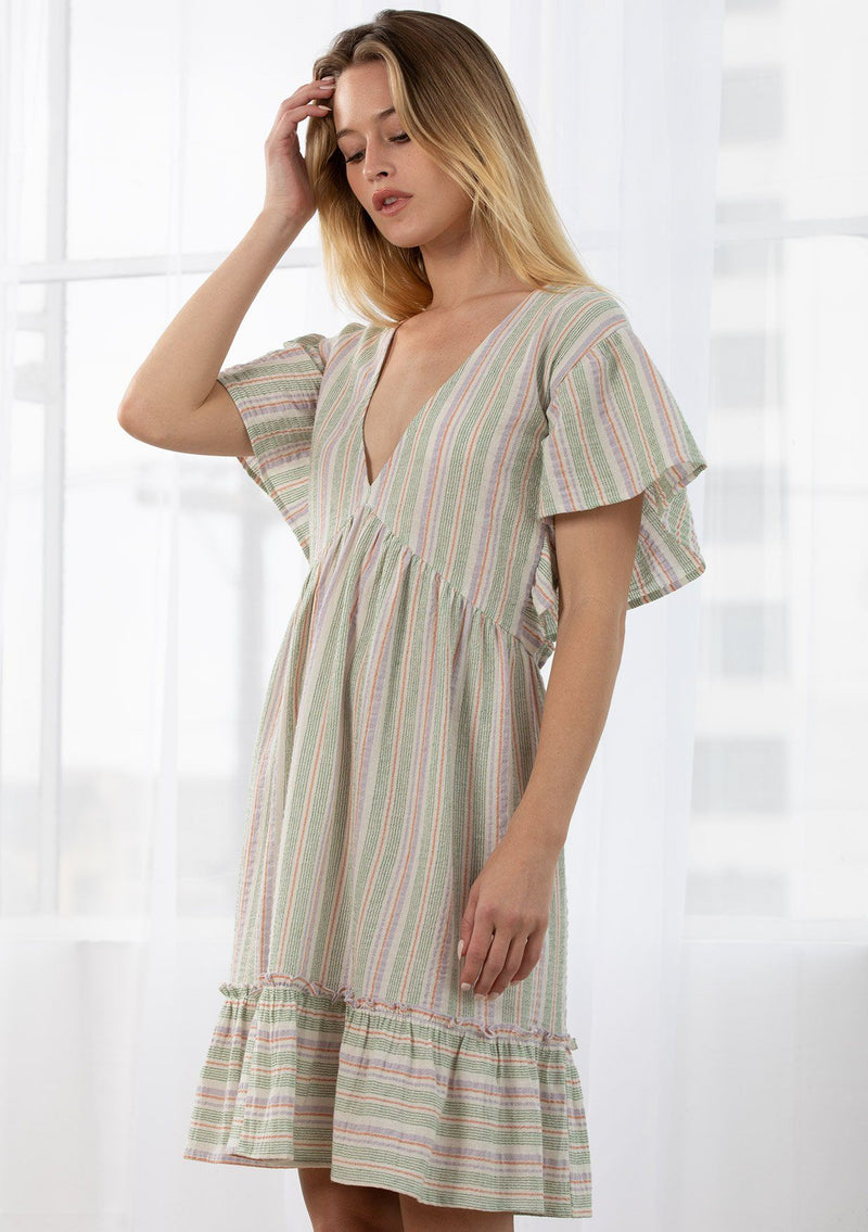[Color: Lilac/Green] A blond model wearing a relaxed, oversize baby doll mini dress in a multi color yarn dye stripe. Featuring short flutter sleeves, a tiered ruffle trimmed skirt, and a plunging v neckline. 