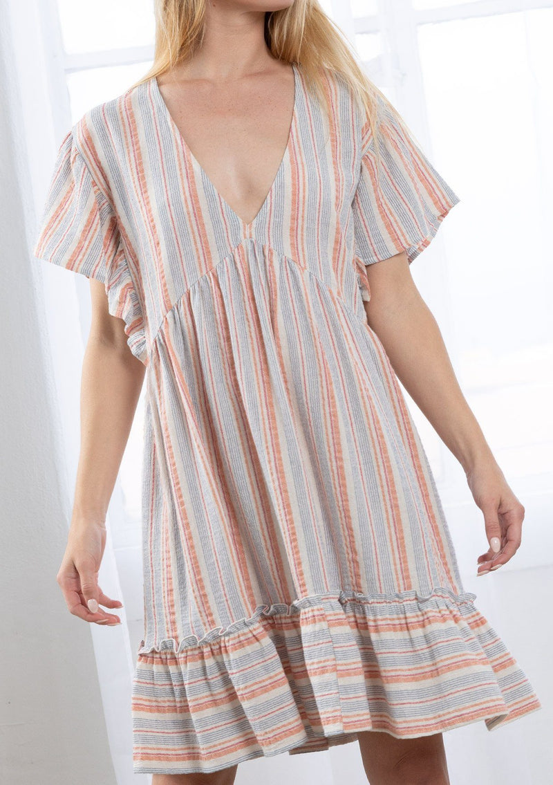 [Color: Coral/Navy] A blond model wearing a relaxed, oversize baby doll mini dress in a multi color yarn dye stripe. Featuring short flutter sleeves, a tiered ruffle trimmed skirt, and a plunging v neckline. 