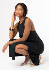 [Color: Black] A side facing image of a black model sitting, wearing a soft and stretchy sleeveless black maxi dress. With a round crew neckline, a sexy side slit, a racer back with a knot detail, and a relaxed fit.