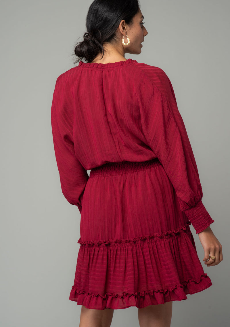 [Color: Burgundy] A back facing image of a burgundy red bohemian mini dress in a textured shadow stripe. With voluminous long dolman sleeves and smocked elastic wrist cuffs, a split v neckline with tassel ties, a ruffle trimmed tiered skirt, and a smocked elastic waist. 
