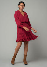 [Color: Burgundy] A full body front facing image of a burgundy red bohemian mini dress in a textured shadow stripe. With voluminous long dolman sleeves and smocked elastic wrist cuffs, a split v neckline with tassel ties, a ruffle trimmed tiered skirt, and a smocked elastic waist. 