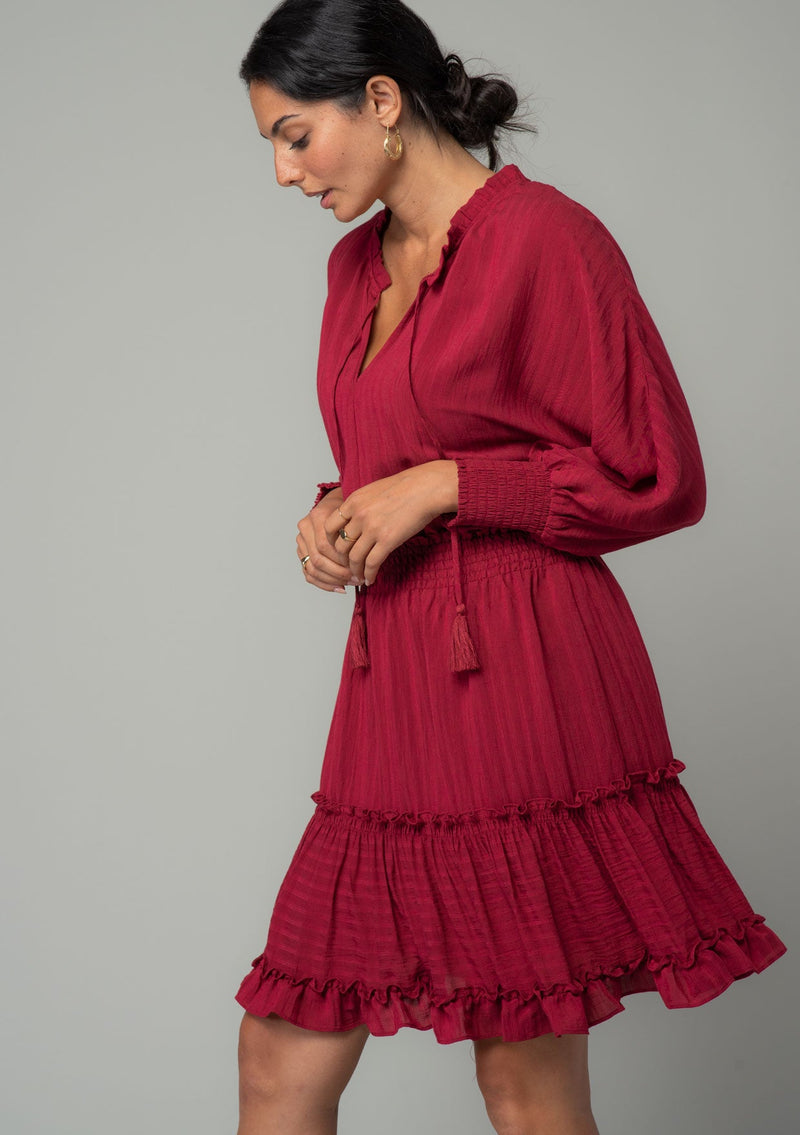 [Color: Burgundy] A side facing image of a burgundy red bohemian mini dress in a textured shadow stripe. With voluminous long dolman sleeves and smocked elastic wrist cuffs, a split v neckline with tassel ties, a ruffle trimmed tiered skirt, and a smocked elastic waist. 