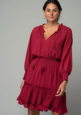 [Color: Burgundy] A half body front facing image of a burgundy red bohemian mini dress in a textured shadow stripe. With voluminous long dolman sleeves and smocked elastic wrist cuffs, a split v neckline with tassel ties, a ruffle trimmed tiered skirt, and a smocked elastic waist. 