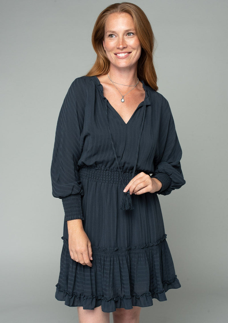 [Color: Charcoal] A half body front facing image of a red headed model wearing a dark grey bohemian mini dress in a textured shadow stripe. With voluminous long dolman sleeves and smocked elastic wrist cuffs, a split v neckline with tassel ties, a ruffle trimmed tiered skirt, and a smocked elastic waist.