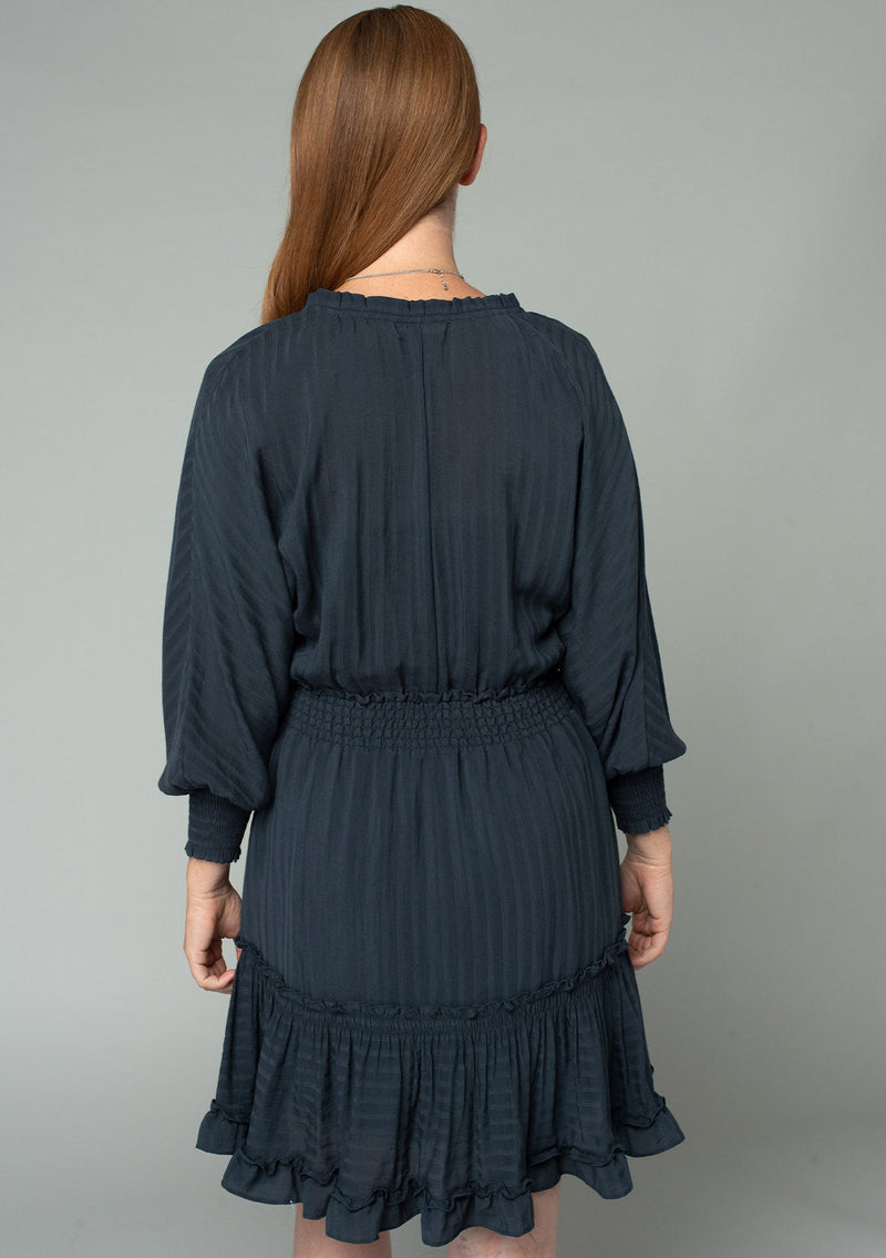 [Color: Charcoal] A back facing image of a red headed model wearing a dark grey bohemian mini dress in a textured shadow stripe. With voluminous long dolman sleeves and smocked elastic wrist cuffs, a split v neckline with tassel ties, a ruffle trimmed tiered skirt, and a smocked elastic waist.