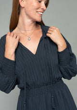 [Color: Charcoal] A close up front facing image of a red headed model wearing a dark grey bohemian mini dress in a textured shadow stripe. With voluminous long dolman sleeves and smocked elastic wrist cuffs, a split v neckline with tassel ties, a ruffle trimmed tiered skirt, and a smocked elastic waist.