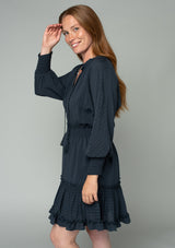 [Color: Charcoal] A side facing image of a red headed model wearing a dark grey bohemian mini dress in a textured shadow stripe. With voluminous long dolman sleeves and smocked elastic wrist cuffs, a split v neckline with tassel ties, a ruffle trimmed tiered skirt, and a smocked elastic waist.