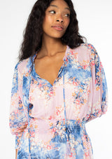 [Color: Lavender/Denim Blue] A close up front facing image of a black model wearing a bohemian lavender purple and blue floral print chiffon mini dress with long sleeves and tassel neck ties. 