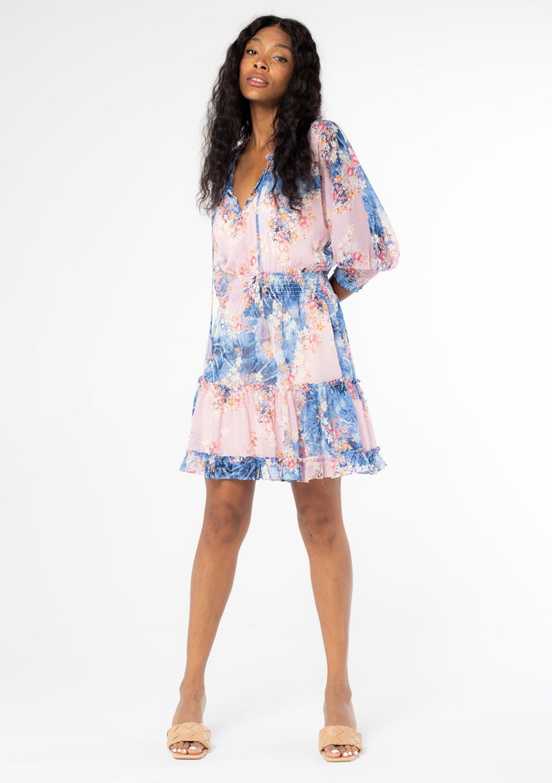 [Color: Lavender/Denim Blue] A full body front facing image of a black model wearing a bohemian lavender purple and blue floral print chiffon mini dress with long sleeves and tassel neck ties. 