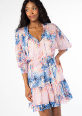 [Color: Lavender/Denim Blue] A half body front facing image of a black model wearing a bohemian lavender purple and blue floral print chiffon mini dress with long sleeves and tassel neck ties. 