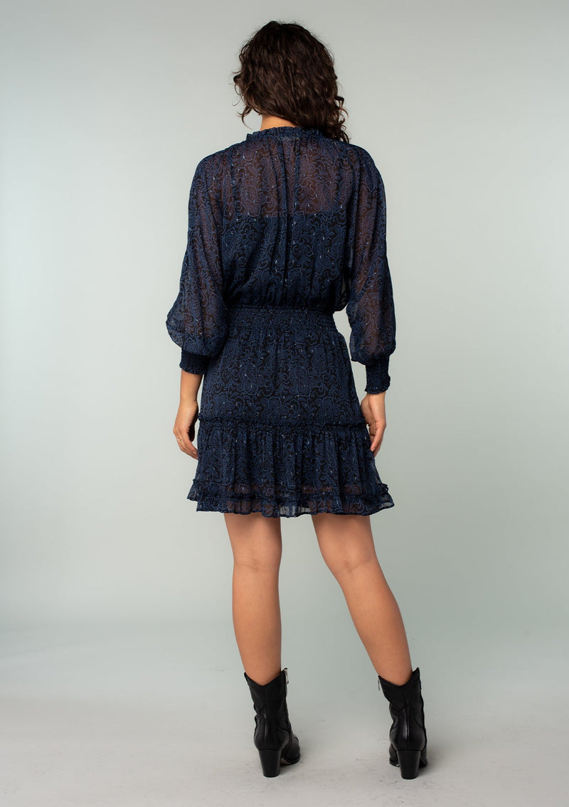 [Color: Black/Blue] A back facing image of a brunette model wearing a black and navy blue paisley print chiffon mini dress with silver clip dot thread accents throughout. With long sheer sleeves, a smocked elastic waist, a split ruffled neckline with tassel ties, and a flowy ruffle trimmed tiered mini skirt.