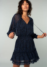 [Color: Black/Blue] A front facing image of a brunette model wearing a black and navy blue paisley print chiffon mini dress with silver clip dot thread accents throughout. With long sheer sleeves, a smocked elastic waist, a split ruffled neckline with tassel ties, and a flowy ruffle trimmed tiered mini skirt.