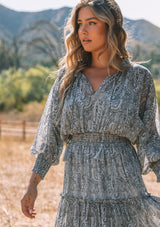 [Color: Grey/Natural] A close up front facing image of a blonde model wearing a best selling bohemian mini dress in sheer chiffon, designed in a grey and natural paisley print. With long sleeves, smocked elastic wrist cuffs, a smocked elastic waist, ruffle trimmed tiered skirt, and tassel tie neckline. 