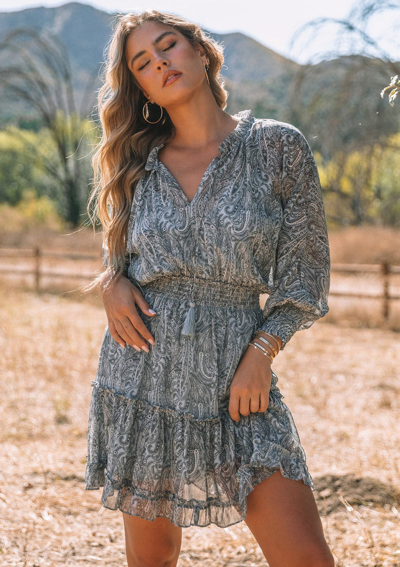 [Color: Grey/Natural] A front facing image of a blonde model wearing a best selling bohemian mini dress in sheer chiffon, designed in a grey and natural paisley print. With long sleeves, smocked elastic wrist cuffs, a smocked elastic waist, ruffle trimmed tiered skirt, and tassel tie neckline. 