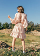 [Color: Sorbet] A woman standing in a field outside wearing a bohemian chic mini dress with long billowy volume sleeves, a beautiful open back with tassel tie details, and made with a soft swiss dot fabrication.