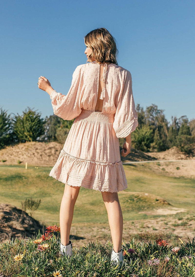 [Color: Sorbet] A woman standing in a field outside wearing a bohemian chic mini dress with long billowy volume sleeves, a beautiful open back with tassel tie details, and made with a soft swiss dot fabrication.