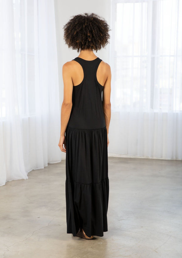 [Color: Black] A model wearing a timeless cotton blend sleeveless black maxi dress. With a dropped waist, tiered skirt, scooped neckline, and racer back detail. 