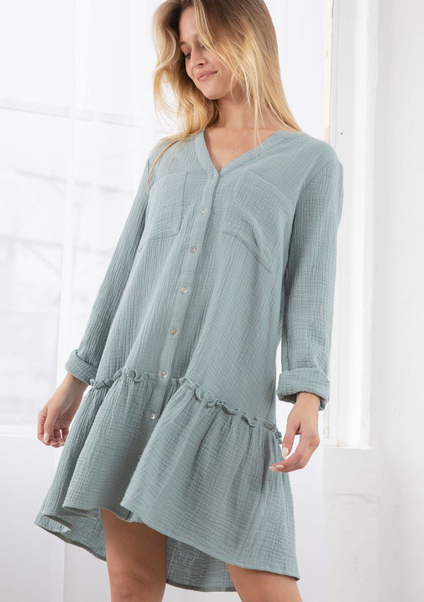 [Color: Dusty Teal] A blond model wearing a cotton gauze button front mini dress. With long rolled sleeves and a button tab closure, a ruffle trimmed dropped waist, and a banded collar with a v neckline.