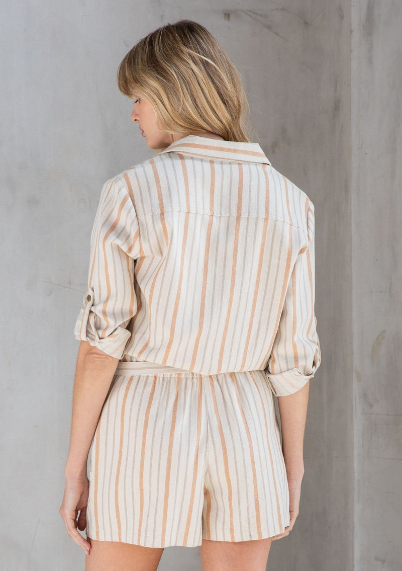[Color: Natural/Terracotta] A model wearing a linen blend twill short romper in a terracotta yarn dye stripe. With long rolled tab sleeves and a tie waist belt.