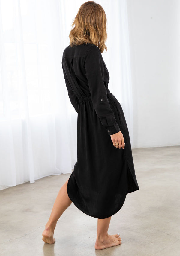 [Color: Black] A model wearing a classic black mid length shirt dress. With a button up front, a collared neckline, front pockets, a drawstring waist, and side pockets. 