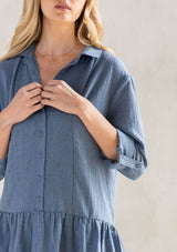 [Color: Denim] A model wearing a blue baby doll mini dress with a button front, a classic collared neckline, long rolled sleeves with a button tab closure, and a tiered skirt.