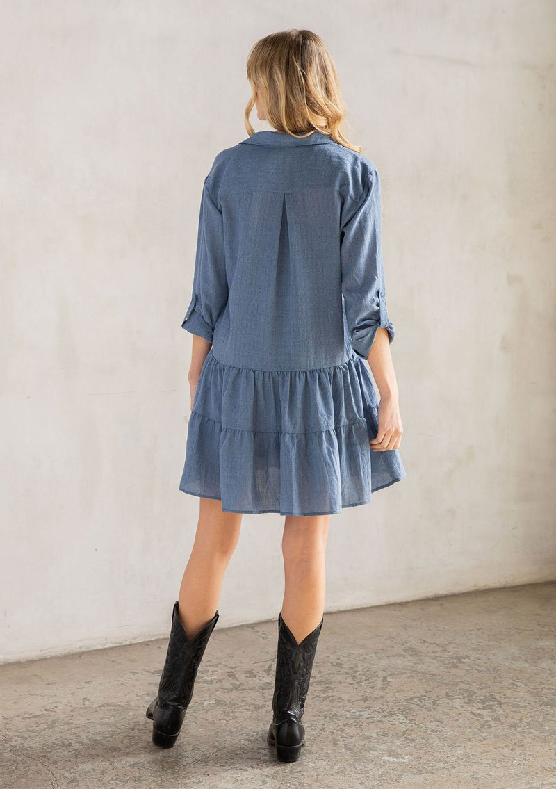 [Color: Denim] A model wearing a blue baby doll mini dress with a button front, a classic collared neckline, long rolled sleeves with a button tab closure, and a tiered skirt.
