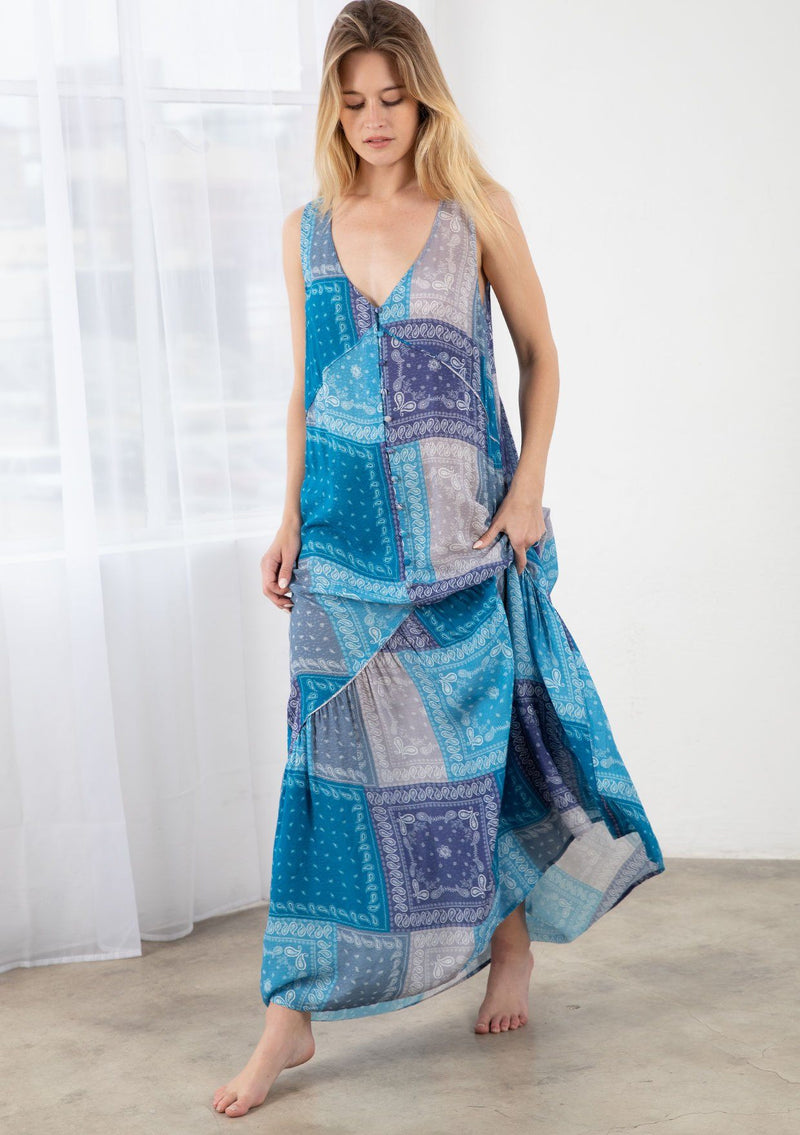 [Color: Purple/Blue] A model wearing a purple and blue paisley bandana patchwork print maxi dress. With a self covered button front, a tiered skirt, a v neckline, thicker tank top straps, and a loose flowy fit. 