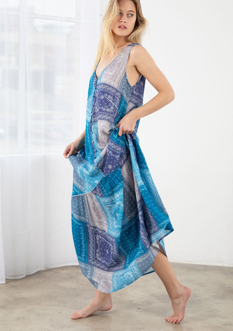 [Color: Purple/Blue] A model wearing a purple and blue paisley bandana patchwork print maxi dress. With a self covered button front, a tiered skirt, a v neckline, thicker tank top straps, and a loose flowy fit. 