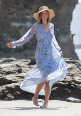[Color: Light Blue] A woman on the beach wearing a dreamy vintage floral maxi dress. Featuring long voluminous sheer sleeves with a flounce wrist cuff, a button front, and an empire waist.