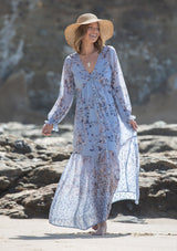 [Color: Light Blue] A woman on the beach wearing a dreamy vintage floral maxi dress. Featuring long voluminous sheer sleeves with a flounce wrist cuff, a button front, and an empire waist.
