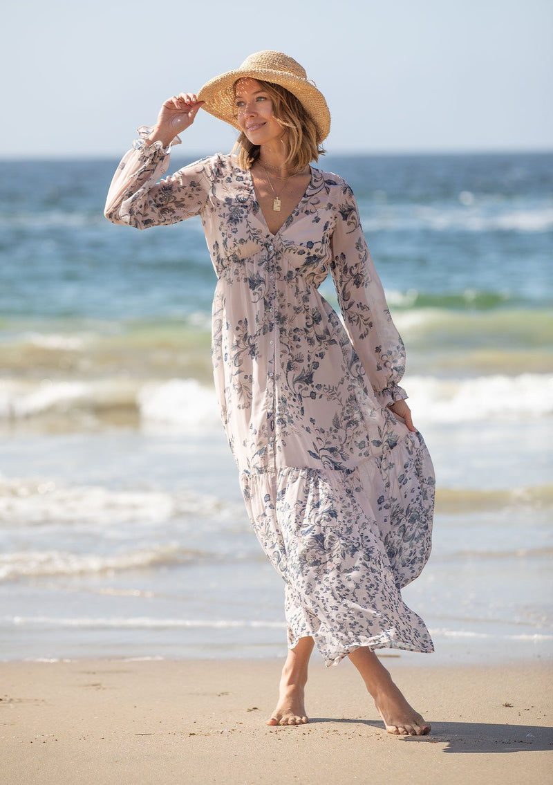 [Color: Light Peach] A woman on the beach wearing a dreamy vintage floral maxi dress. Featuring long voluminous sheer sleeves with a flounce wrist cuff, a button front, and an empire waist.