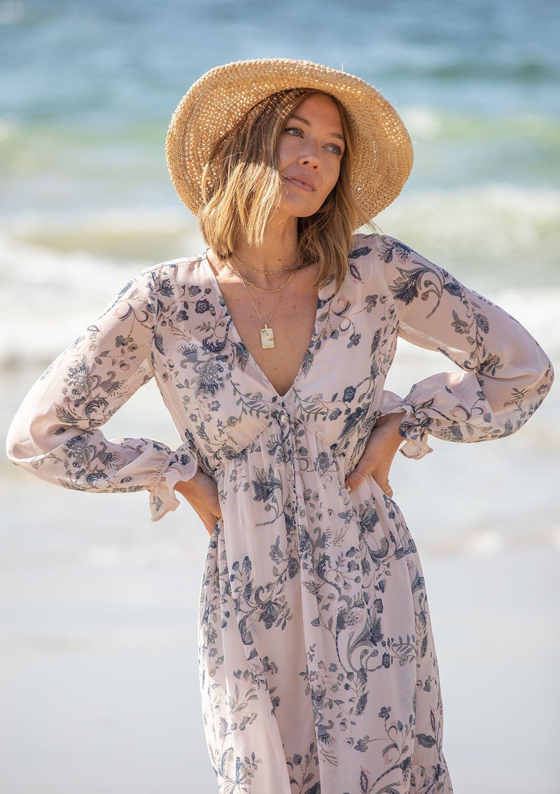 [Color: Light Peach] A woman on the beach wearing a dreamy vintage floral maxi dress. Featuring long voluminous sheer sleeves with a flounce wrist cuff, a button front, and an empire waist.