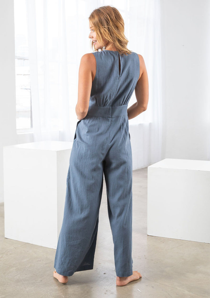 [Color: Denim Blue] A model wearing linen blend full length jumpsuit. With a wide leg, side pockets, a v neckline, and a back zip closure. 