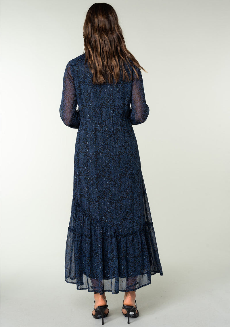 [Color: Black/Blue] A full body back facing image of a brunette model wearing a best selling navy blue chiffon maxi dress with black paisley print and light catching metallic clip dot details. Perfect for special occasions, with sheer long sleeves, a v neckline, an empire waist with drawstring tassel tie detail, and an asymmetric ruffled trimmed tiered skirt. 