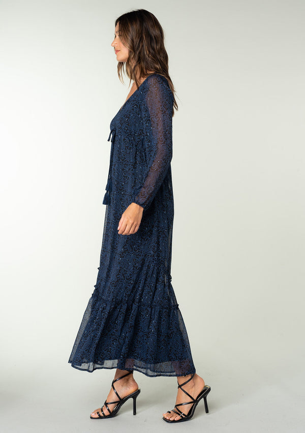 [Color: Black/Blue] A full body side facing image of a brunette model wearing a best selling navy blue chiffon maxi dress with black paisley print and light catching metallic clip dot details. Perfect for special occasions, with sheer long sleeves, a v neckline, an empire waist with drawstring tassel tie detail, and an asymmetric ruffled trimmed tiered skirt. 