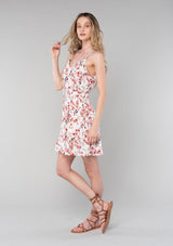 [Color: Natural/Rose] A side facing image of a blonde model wearing a bohemian spring mini dress in a rose pink floral print. With spaghetti straps, a split v neckline with tassel ties, a smocked elastic waist, and a flowy tiered skirt.