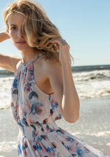 [Color: Ivory/Blue] A close up side facing image of a blonde model at the beach wearing a dreamy sheer floral maxi dress with a braided trimmed halter neckline, a smocked elastic waist, a sexy side slit, and a back keyhole with braided rope tie.
