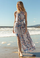[Color: Ivory/Blue] A front facing image of a blonde model at the beach wearing a dreamy sheer floral maxi dress with a braided trimmed halter neckline, a smocked elastic waist, a sexy side slit, and a back keyhole with braided rope tie.