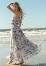 [Color: Ivory/Blue] A side facing image of a blonde model at the beach wearing a dreamy sheer floral maxi dress with a braided trimmed halter neckline, a smocked elastic waist, a sexy side slit, and a back keyhole with braided rope tie.