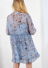[Color: Light Blue] A blond model wearing a sheer floral print mini dress. Featuring three quarter length sleeves, a button front, and a ruffle trimmed waist.