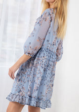 [Color: Light Blue] A blond model wearing a sheer floral print mini dress. Featuring three quarter length sleeves, a button front, and a ruffle trimmed waist.