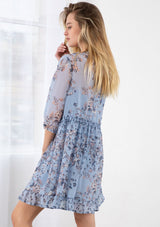 [Color: Light Blue] A blond model wearing a sheer floral print mini dress. Featuring three quarter length sleeves, a button front, and a ruffle trimmed waist.