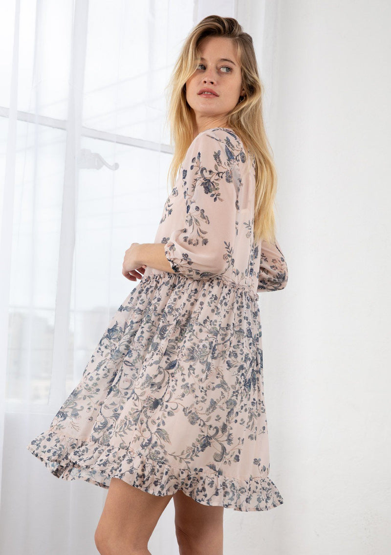 [Color: Light Peach] A blond model wearing a sheer floral print mini dress. Featuring three quarter length sleeves, a button front, and a ruffle trimmed waist.
