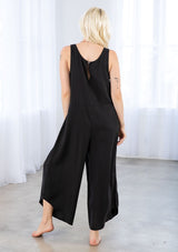 [Color: Black] A model wearing a relaxed oversize sleeveless black jumpsuit. With thick tank top straps, a scooped neckline in front and back, side pockets, and a wide leg with an asymmetric hemline. 