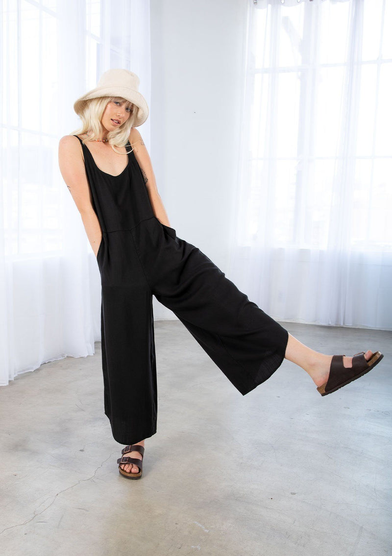 [Color: Black] A model wearing a relaxed oversize sleeveless black jumpsuit. With thick tank top straps, a scooped neckline in front and back, side pockets, and a wide leg with an asymmetric hemline. 