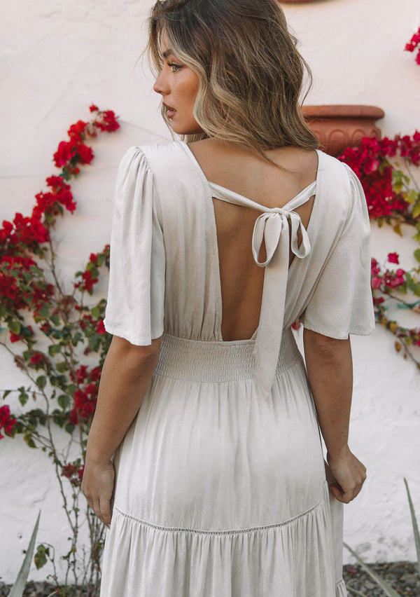 [Color: Vanilla Bean] A model wearing a flowy off white maxi dress in a floral jacquard. With short flutter sleeves and delicate lattice trim.