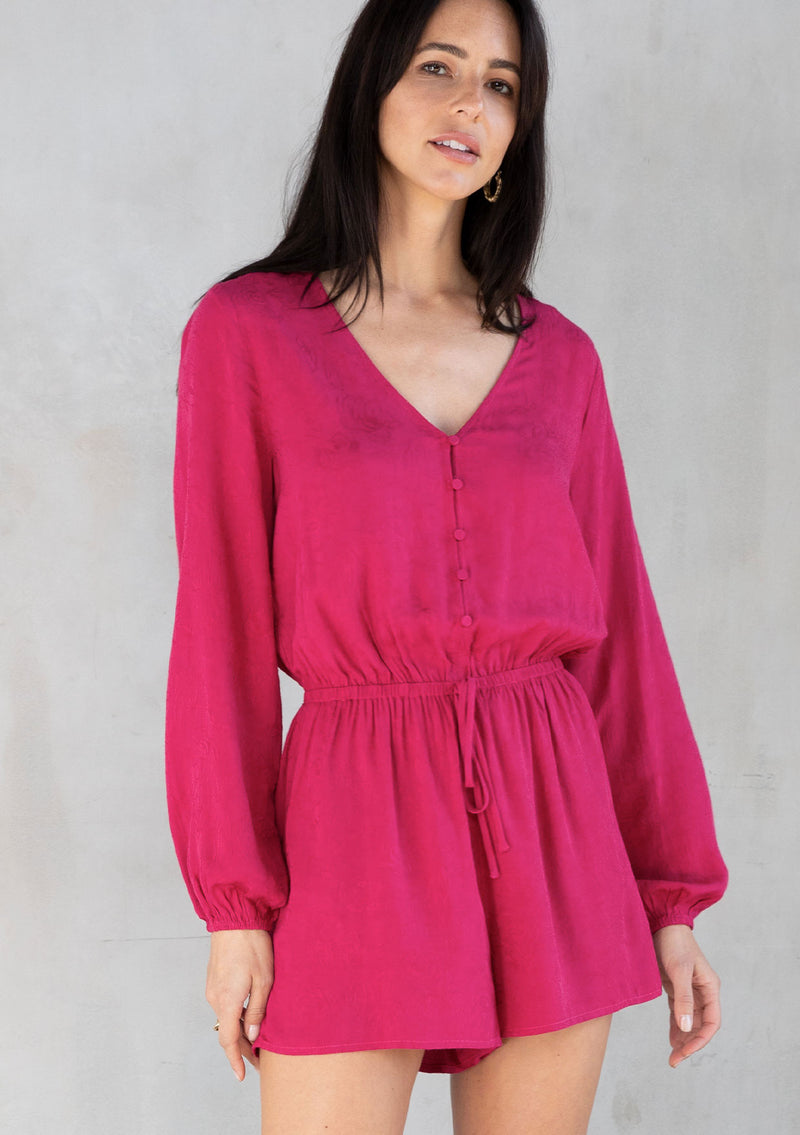 [Color: Mulberry] A model wearing a playful and chic pink short romper in rose jacquard. With long sleeves, a button up front, and a v neckline.