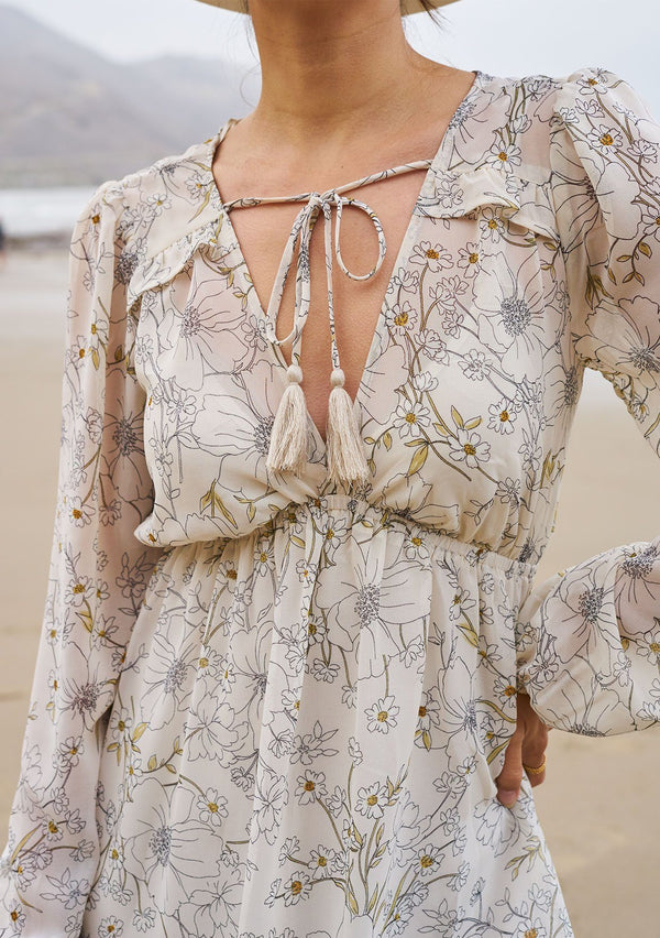 [Color: Natural/Sand] A model wearing a sheer floral mid length dress. With a high low hemline, an empire waistline, long voluminous sleeves, and tassel ties.