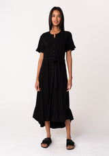 [Color: Black] A front facing image of a model wearing a black mid length shirt dress in a linen blend. Featuring short sleeves with a ruffled trim, a button up front, an adjustable waist tie, and a ruffled hemline.